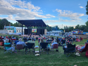 A photo of Littleton residents gathering on the green lawn of the Lakeland Amphitheater. They are sitting in folding chairs in front of the stage. The sky is bright blue with tons of white, puffy clouds.