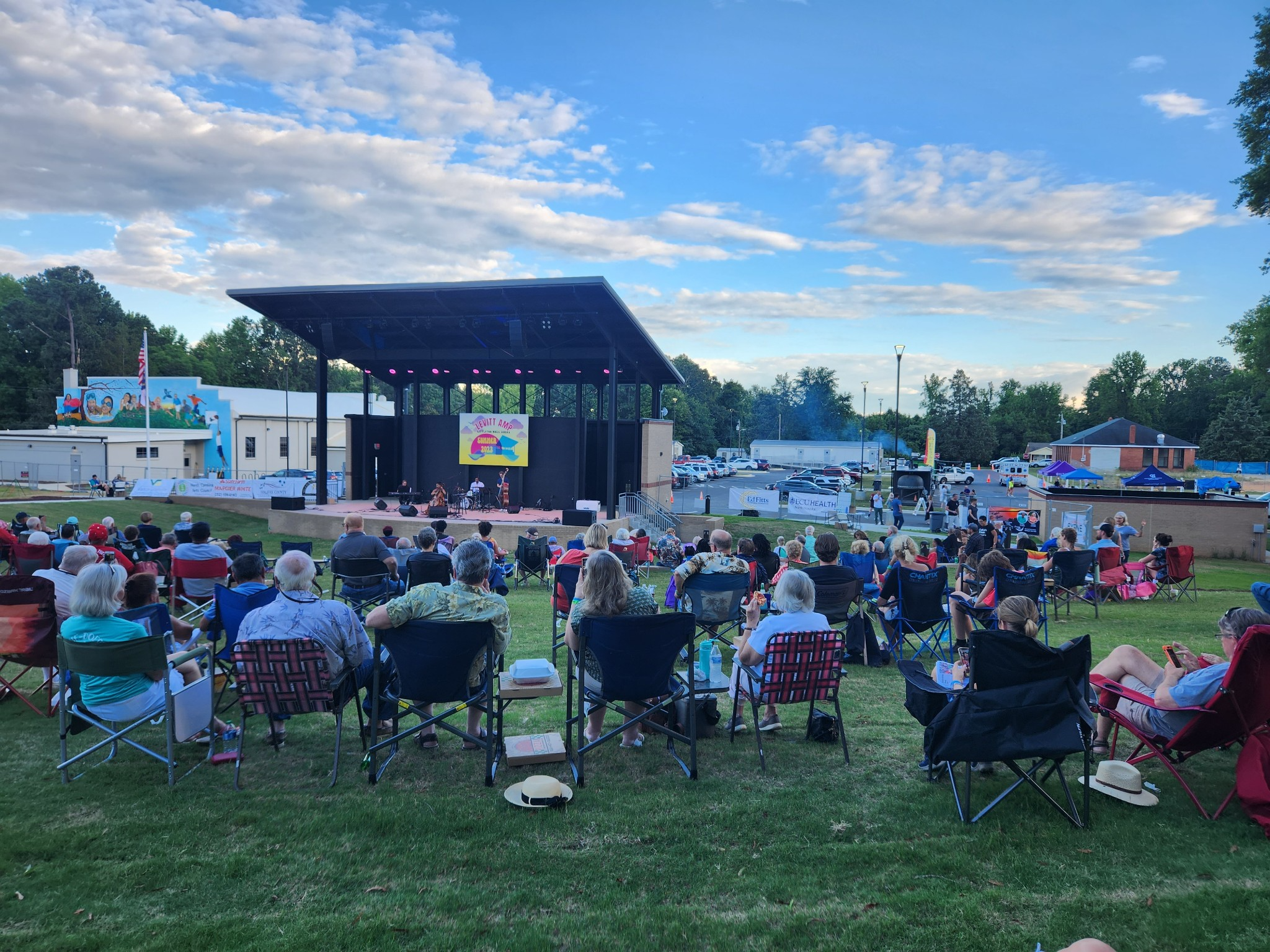 A photo of Littleton residents gathering on the green lawn of the Lakeland Amphitheater. They are sitting in folding chairs in front of the stage. The sky is bright blue with tons of white, puffy clouds.
