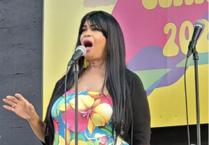 Shown in this image is a Black woman with long dark hair and a floral-printed dress, standing in front of a microphone and singing. 