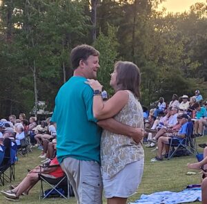 A couple dances happily on a lawn packed with community members. The sky is golden and many lush, tall trees are in the background. 