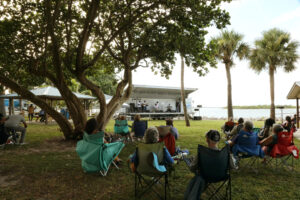 Image shows, from behind, an audience of people sitting in lawn chairs. They are facing a stage off in the distance. It is a sunny day in Florida, with many green palm trees and lush grass. Performers are playing music at the stage. 