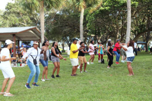 A diverse group of audience members of all ages and ethnicities are shown dancing on a lush, green lawn. An African American woman wearing denim shorts and a white shirt is taking a photo of the dancing folks. There are many palm trees in the background. 