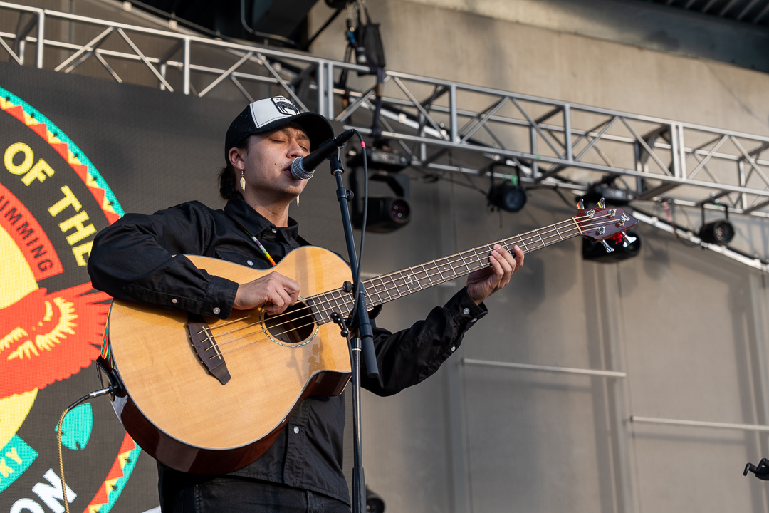 A Native musician, Frank Waln, played an acoustic bass on a stage. He is wearing dangling earrings and a colorful beaded necklace. He is a light-skinned brown man with brown hair and a hat on. His eyes are closed as he plays bass.