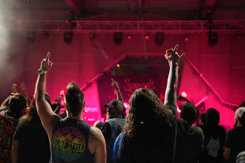 Two folks shown from behind, standing in the crowd at a metal festival. One is holding a beer can with a "rock on" sign, the other has a mohawk with their hands up. The lighting is red and a stage is shown in the background. 