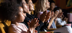 An ethnically diverse row of women and girls sitting in a movie theater row clapping and smiling.